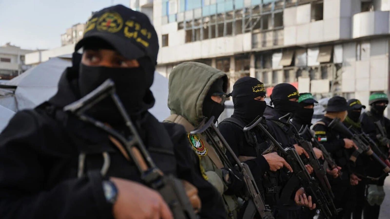 Masked armed fighters in black uniforms with green headbands standing in a row, holding rifles, in front of a damaged building in Gaza.