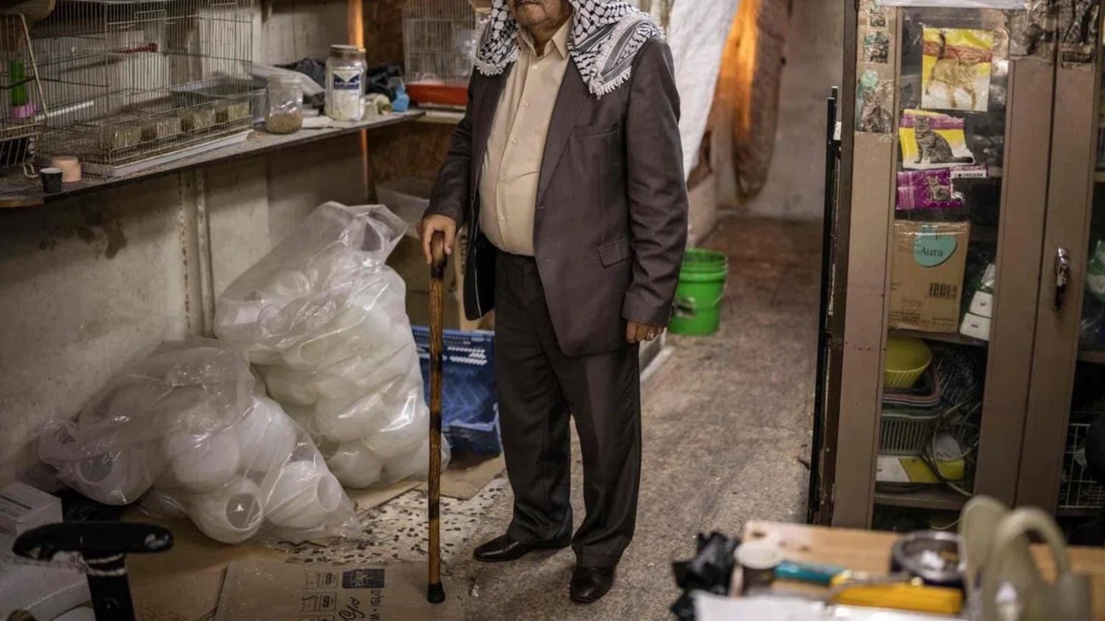An elderly Palestinian man wearing a keffiyeh standing with a cane amid supplies in a cluttered storage room.