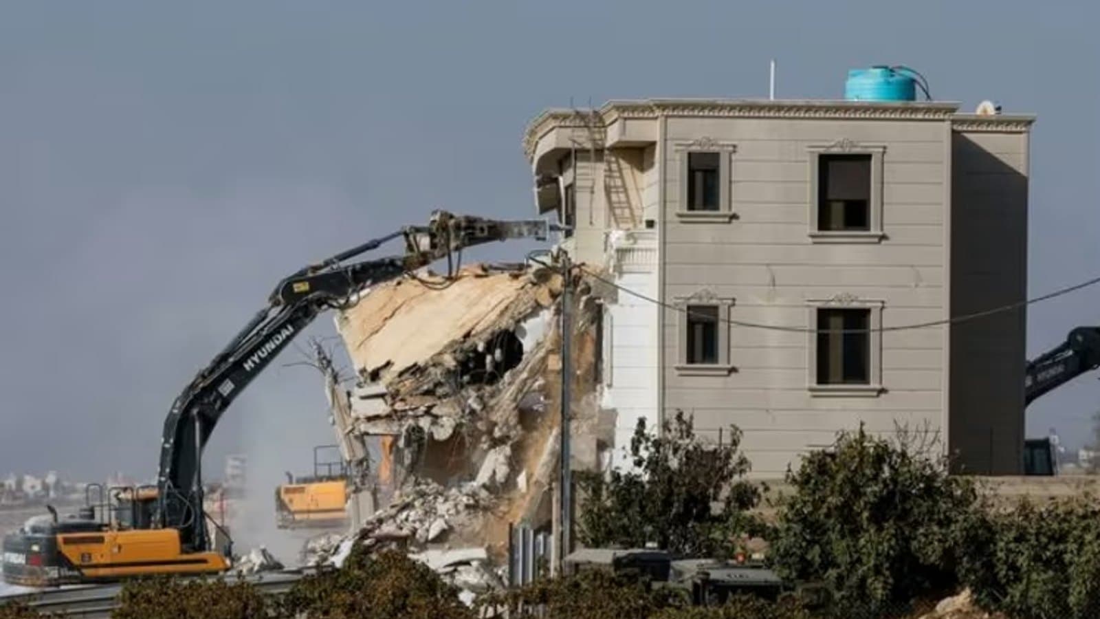 An excavator demolishing a large building in Hebron, with rubble and construction vehicles visible.