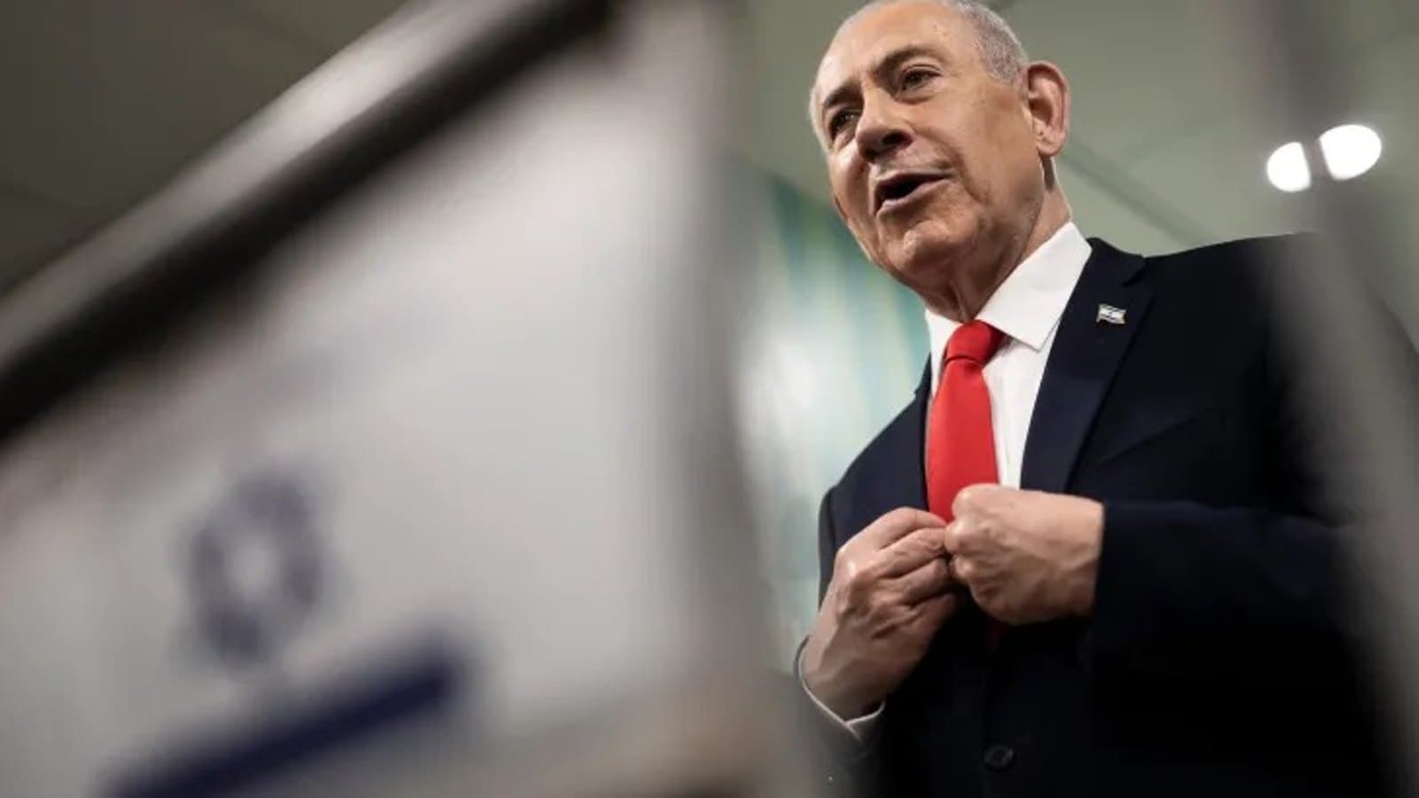 Netanyahu adjusting his jacket while speaking, with an Israeli flag blurred in the background. תצלום ארכיון.
