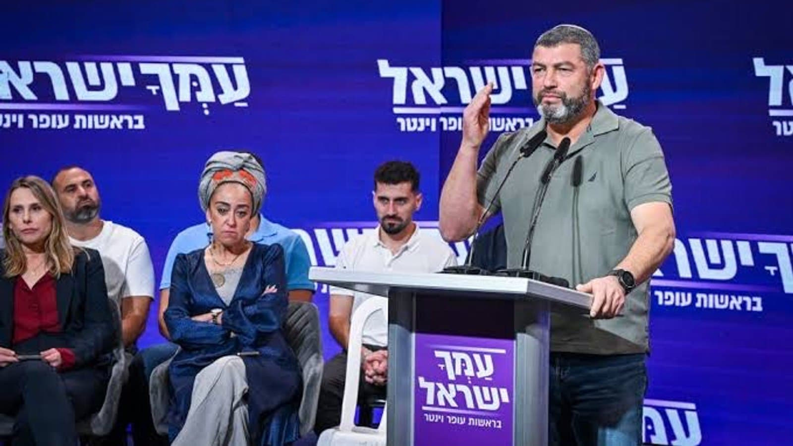 Ofer Winter speaking at a podium during an Amcha Yisrael campaign event, with party colleagues seated behind him.