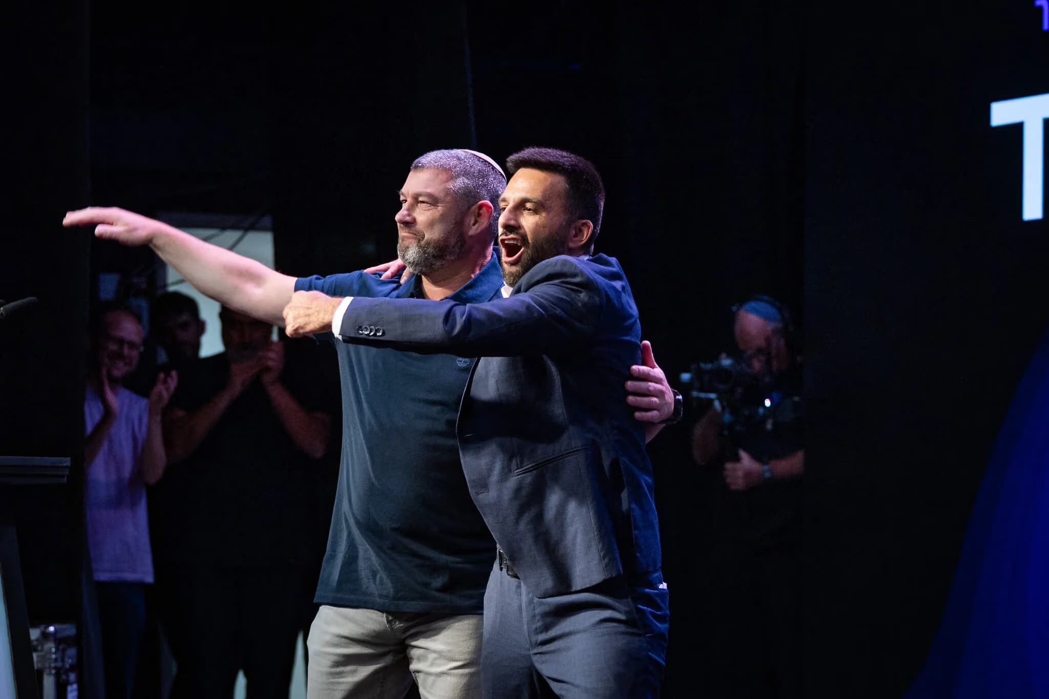 Two men on stage at a political event, one pointing toward the crowd while the other cheers beside him